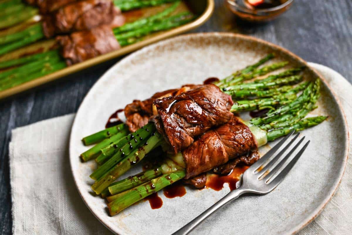 a plate with 3 steak cheese and asparagus rolls topped with a balsamic glaze with a fork