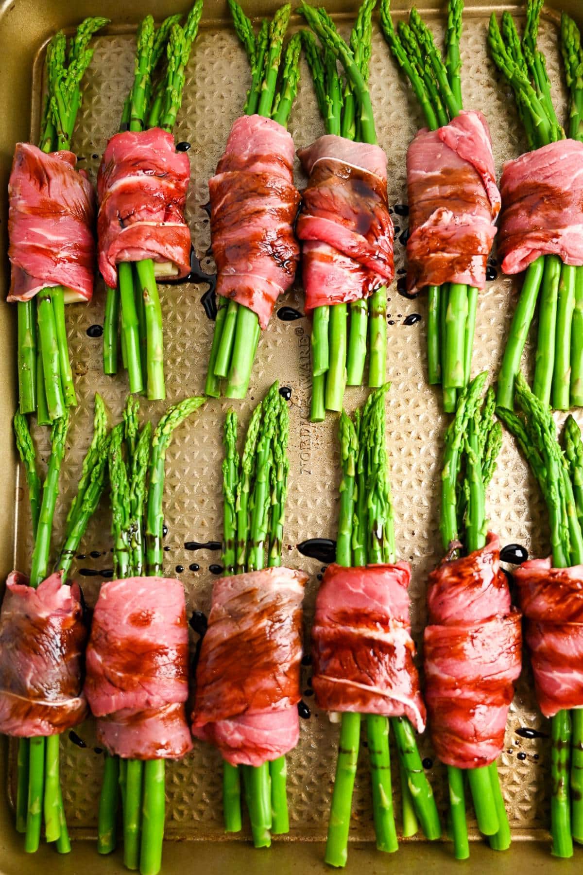 asparagus rolled with steak and cheese on a baking sheet drizzled with balsamic vinegar