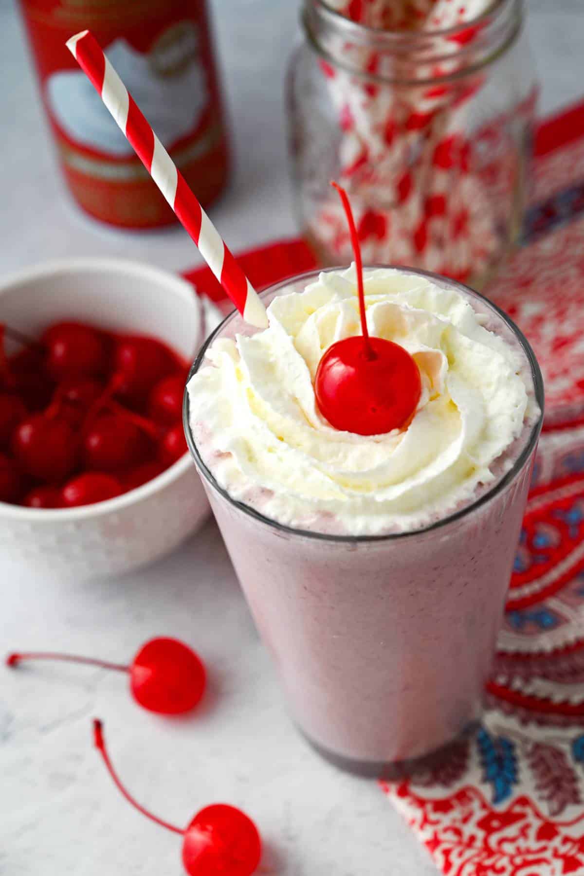 a cherry milkshake surrounded by a bowl of cherries, whipped cream can and straws