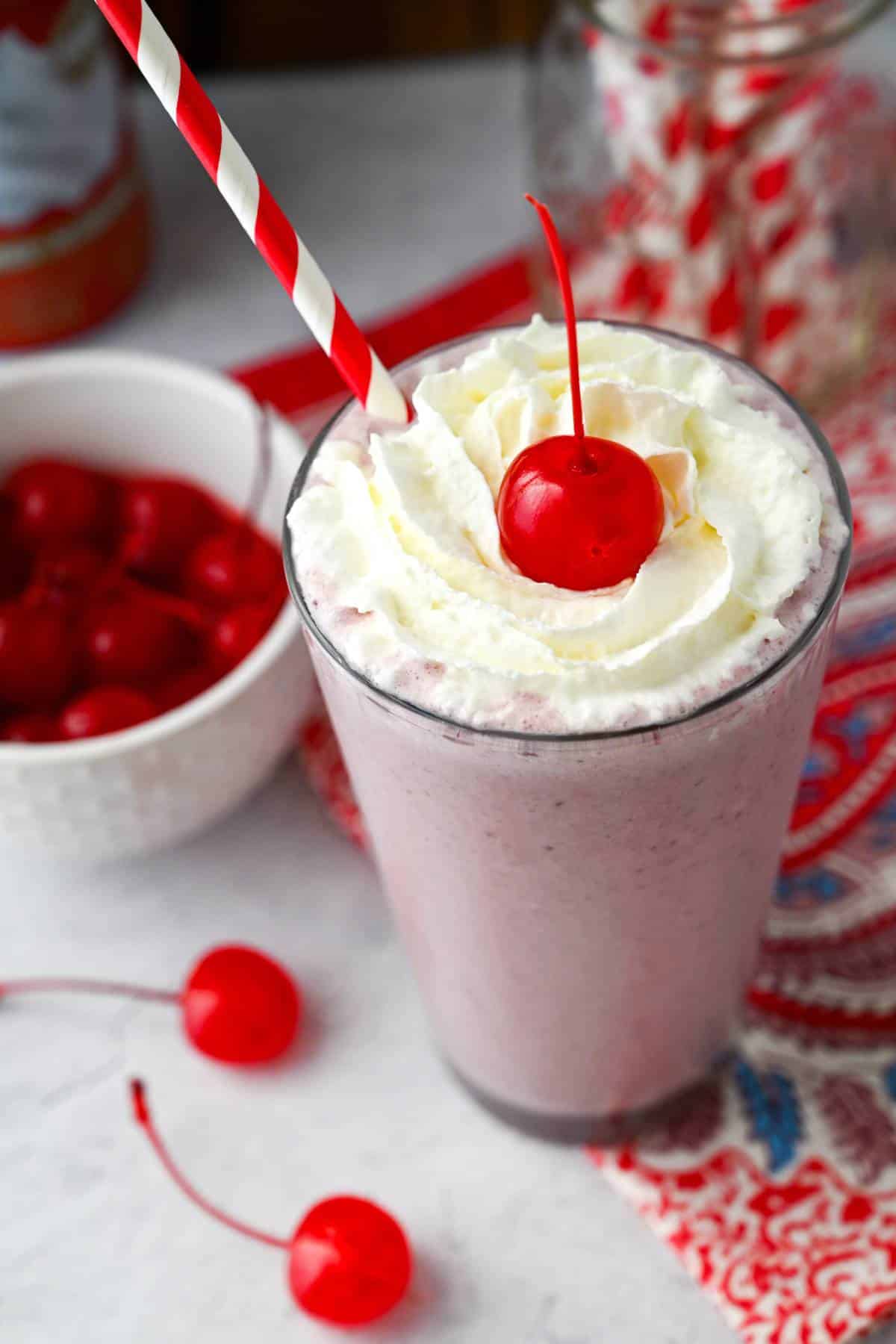 a couple of marachino cherries next to a cherry milkshake in a clear tall glass with a straw