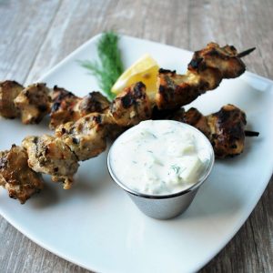 Two chicken skewers on a plate with dipping sauce and fresh herbs