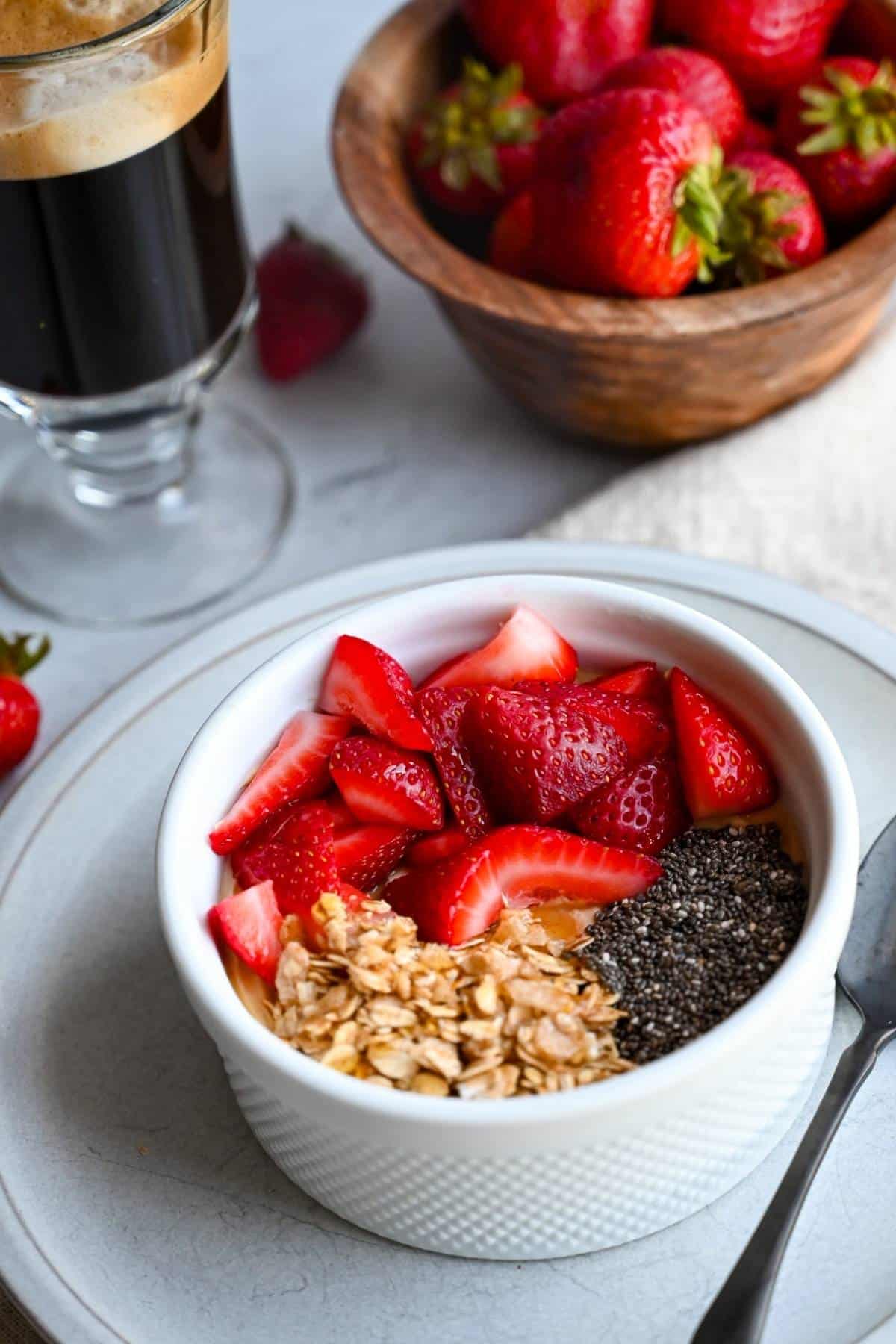 a white dish with high protein yogurt topped with berries, chia seeds, and granola, with a napkin, coffee and berries next to it