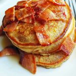 close up of a stack of pancakes on a white plate topped with syrup and chopped bacon