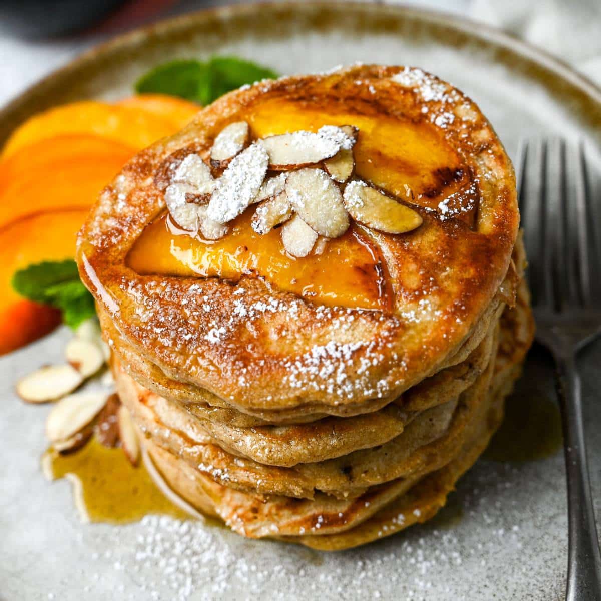 close up of a stack of peach cobbler pancakes on a plate with a fork with mint sprigs and sliced peaches next to it