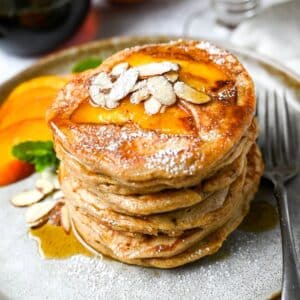 six pancakes stacked on a plate topped with cooked peach slices, almonds, maple syrup and powdered sugar