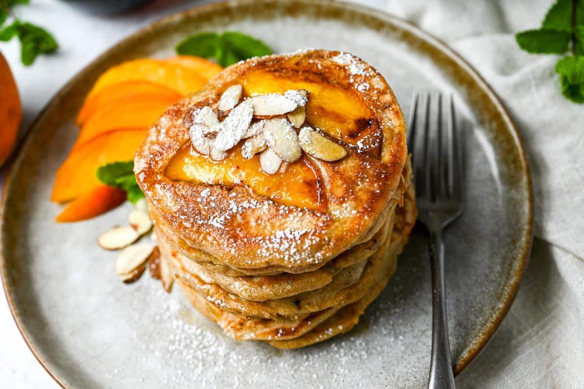 a plate of stacked peach protein pancakes with peach slices, almonds, and powdered sugar with a fork