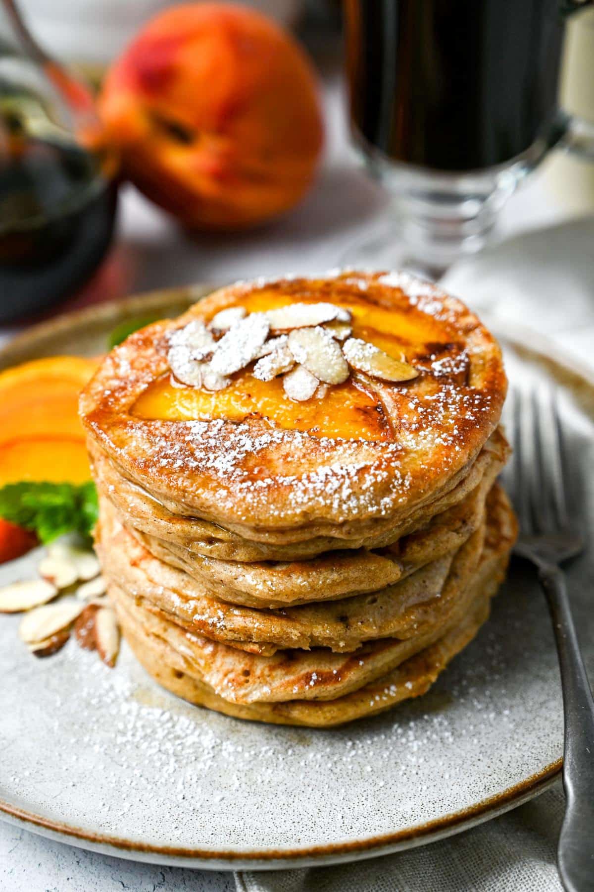 a plate of pancakes with peach slices on top and sliced almonds, maple syrup and powdered sugar with a whole peach and cup of coffee behind it