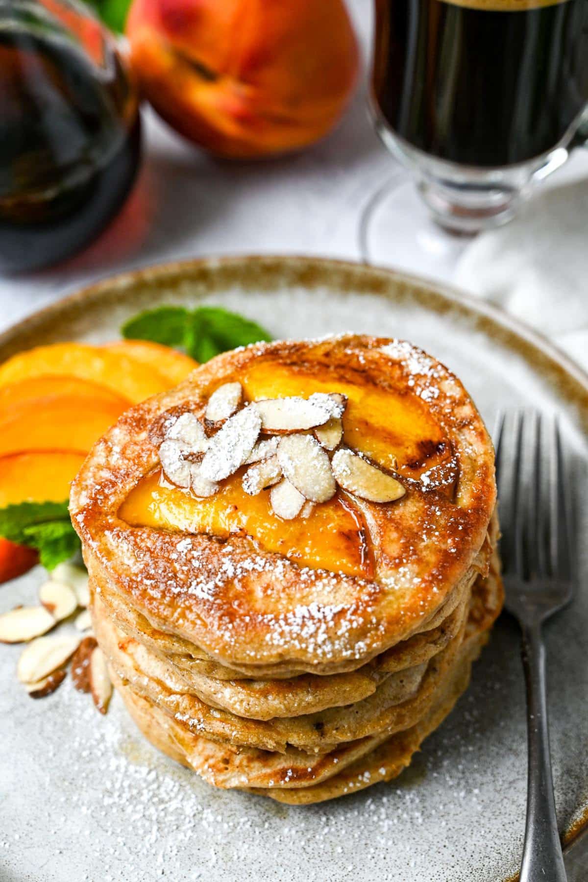 looking down at a stack of peach pancakes with almonds and powdered sugar on a plate with a peach and cup of coffee behind it