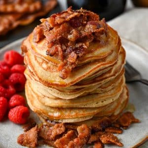 close up of a tall stack of pancakes with chopped candied bacon dripping with maple syrup