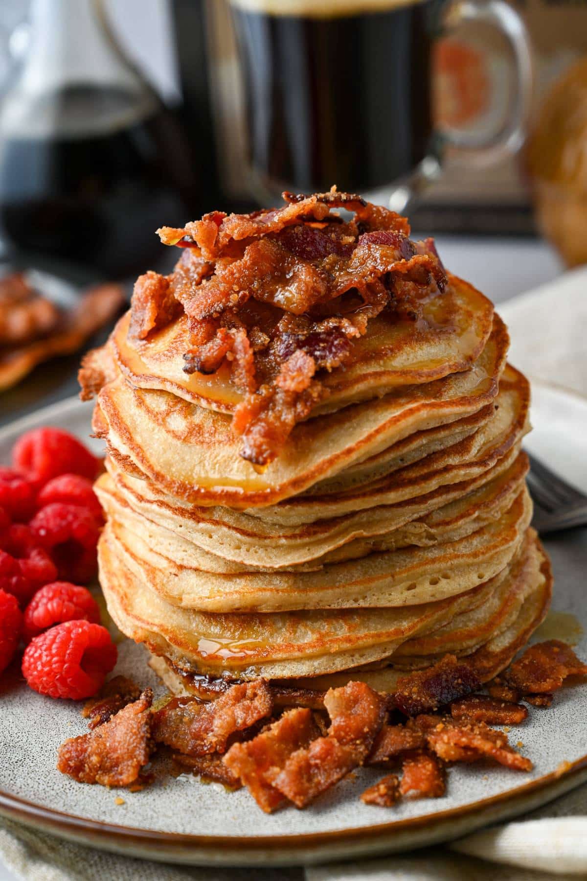 stack of buttermilk protein pancakes topped with candied bacon on a plate with raspberries with a jar of syrup and coffee behind it