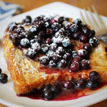 close up of whole grain French toast on a plate topped with wild blueberries and syrup with a fork on the side