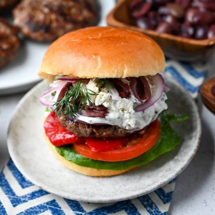 a loaded greek burger with roasted peppers, feta, tzatziki, lettuce, tomatoe, olives, onions, and dill on a brioche bun on a plate