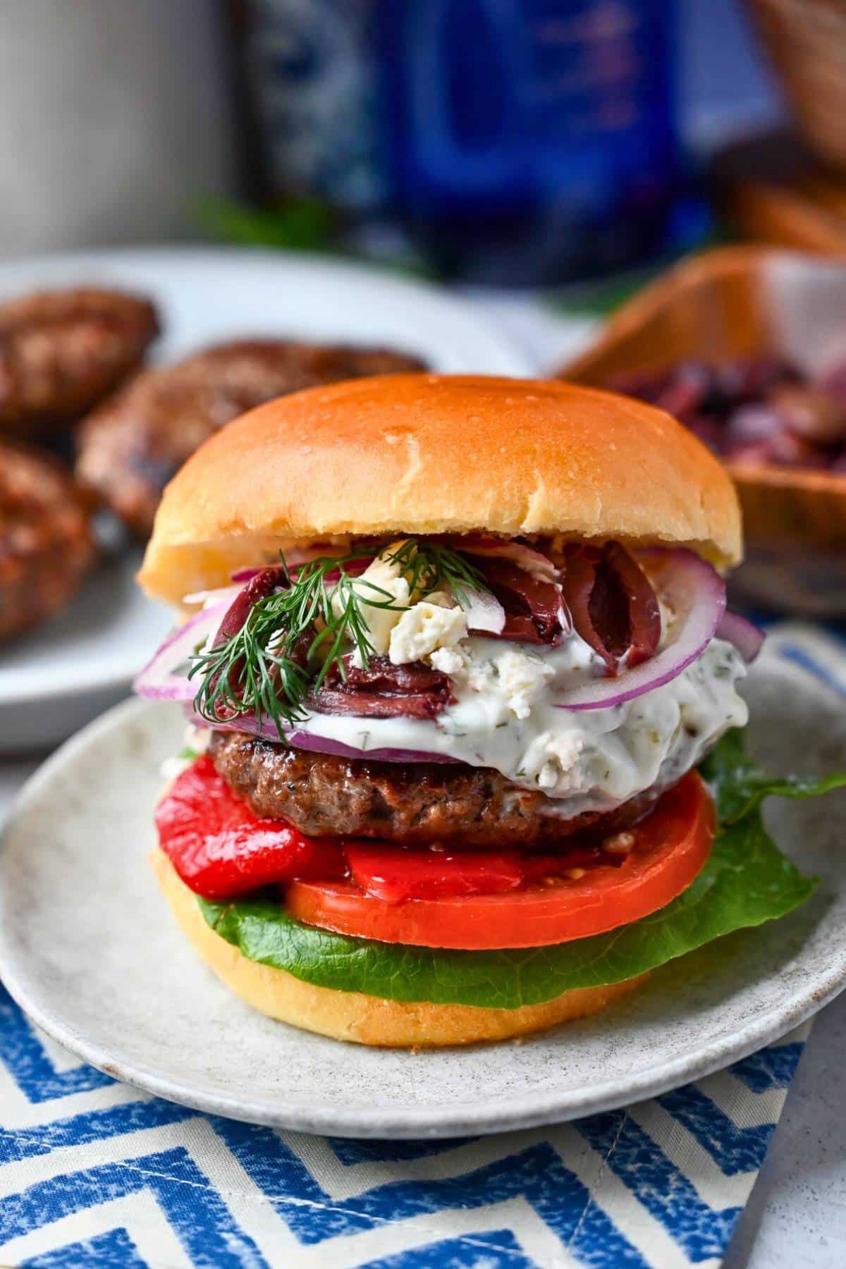 a mediterranean inspired burger with toppings on a plate with a platter of burgers behind it