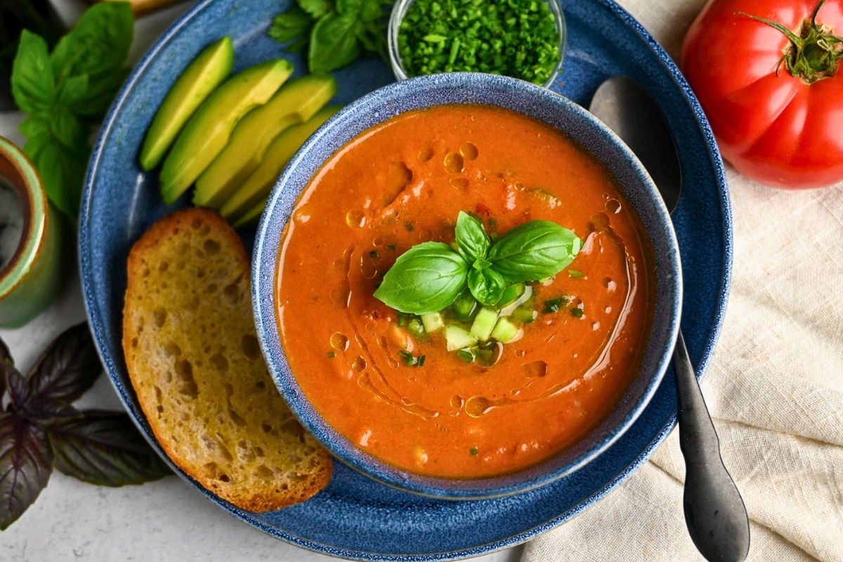 gazpacho garnished with chopped cucumber, chives, and basil on a plate with toasted sourdough and avocado