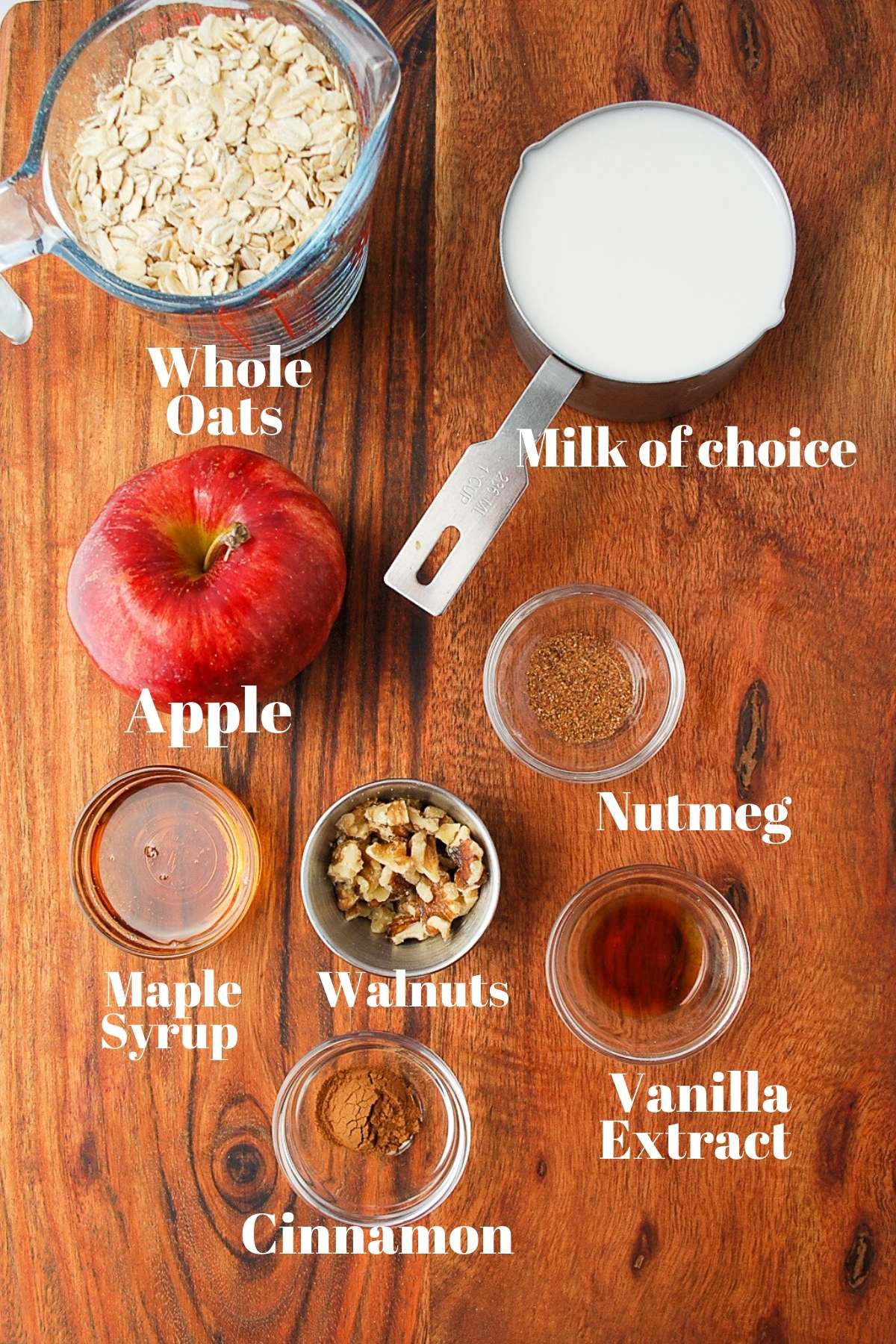 Ingredients for apple pie oats on a wood board: oats, milk, apple, nutmeg, maple syrup, walnuts, vanilla, and cinnamon