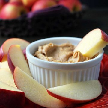 peanut butter dip in a white ramekin with an apple slice dipped inside with slices of apple on a plate around it
