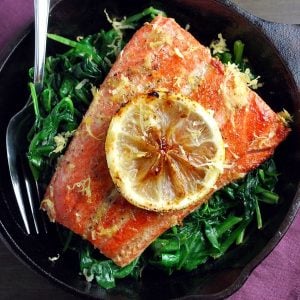 a salmon filet in a cast iron skillet over wilted spinach with a lemon slice on top