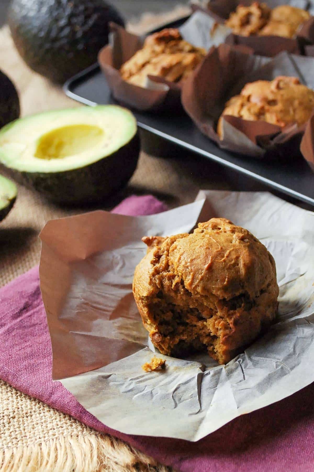 an avocado muffin with a bite taken out of it with a sliced avocado and pan of muffins in the background