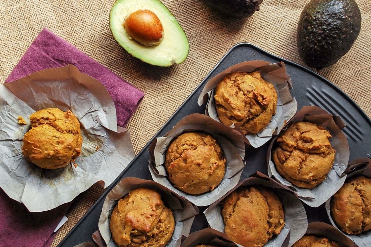 looking down at a pan of muffins with a muffin on top of a parchment liner next to it and a sliced avocado