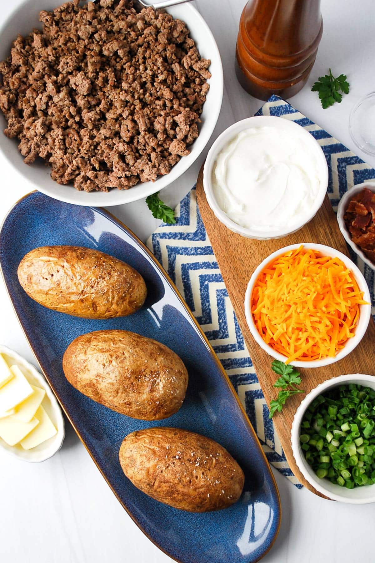 ingredients for a baked potato bar on a table: potatoes, cooked ground beef and sausage, butter, sour cream, cheese, bacon, and green onions