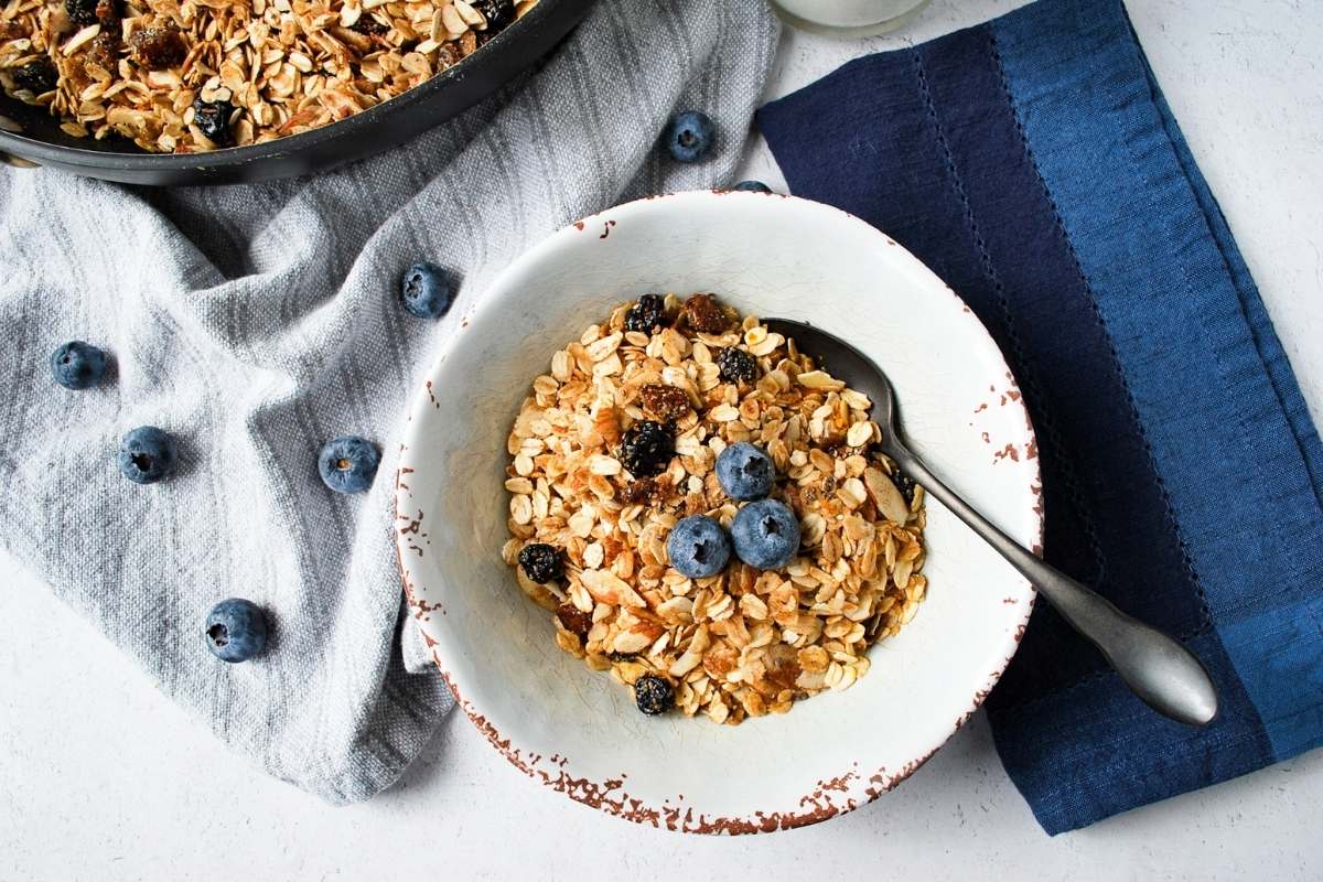granola made with dried blueberries in a bowl surrounded by fresh blueberries and a blue napkin