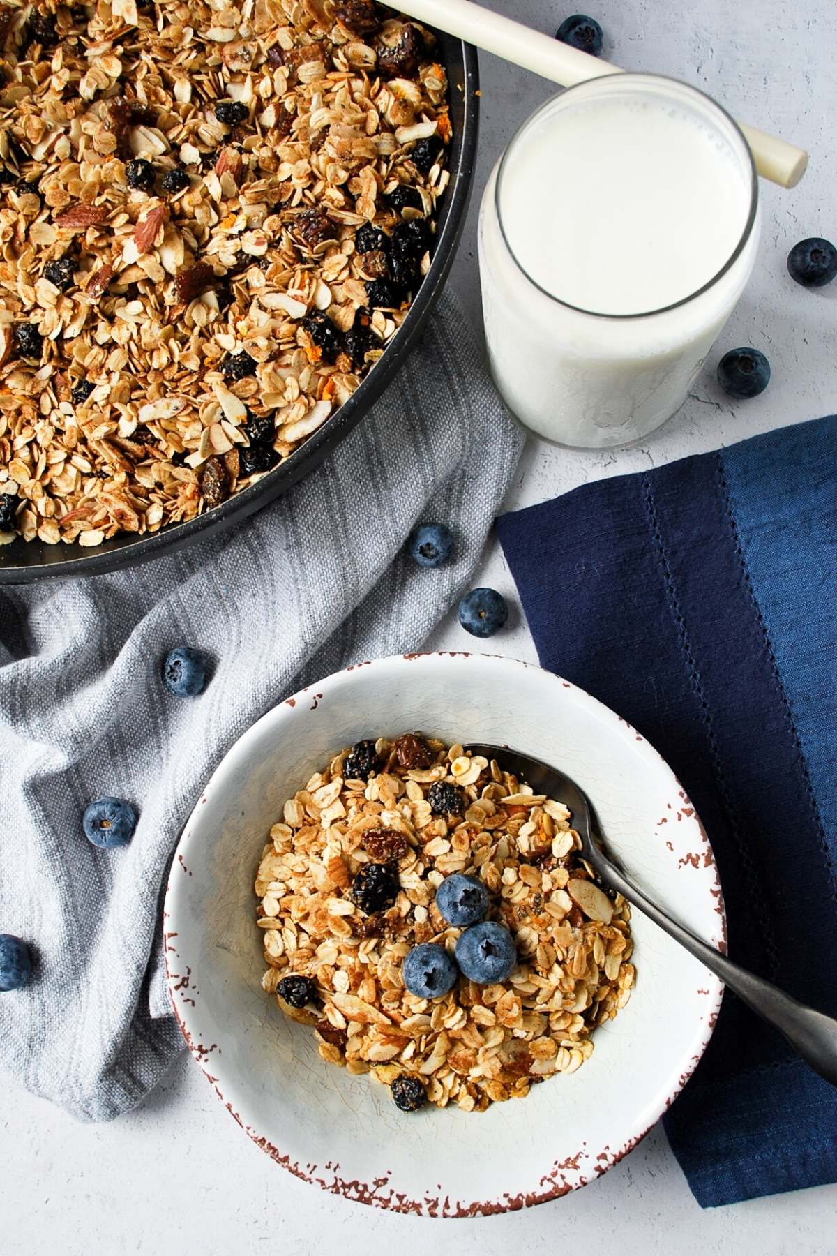 a bowl of blueberry granola next to a skillet of blueberry granola and a glass of milk