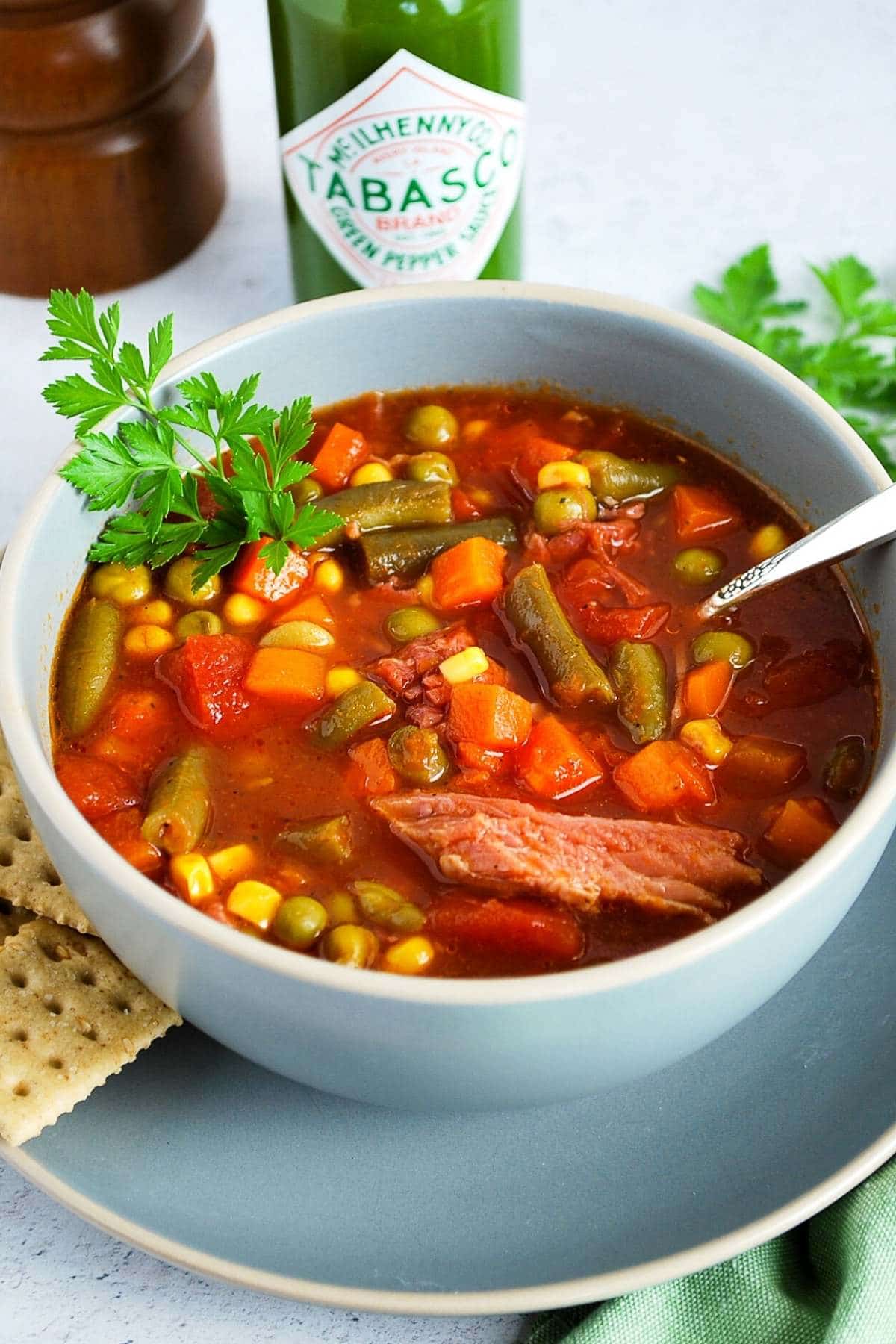 ham vegetable soup in a grey bowl with a spoon and a bottle of Tabasco in the background