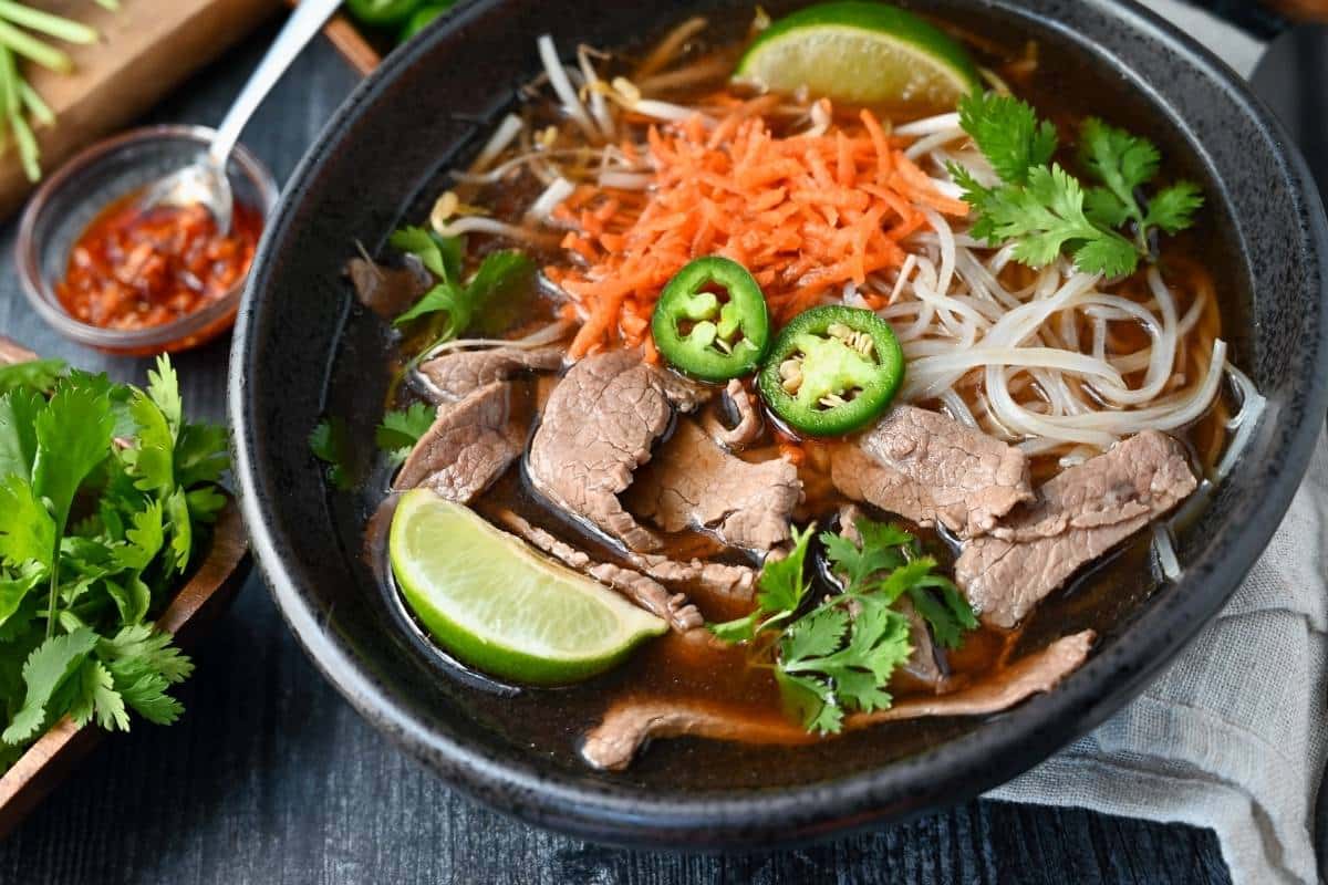 a zoomed in photo of a bowl of flank pho with limes, jalapenos, carrots and bean sprouts