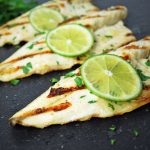 three grilled fish filets on a black cutting board topped with a slice of lime