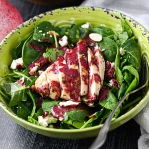close up of a bowl of kale and spinach salad with a grilled chicken breast on top drizzled with blueberry vinaigrette