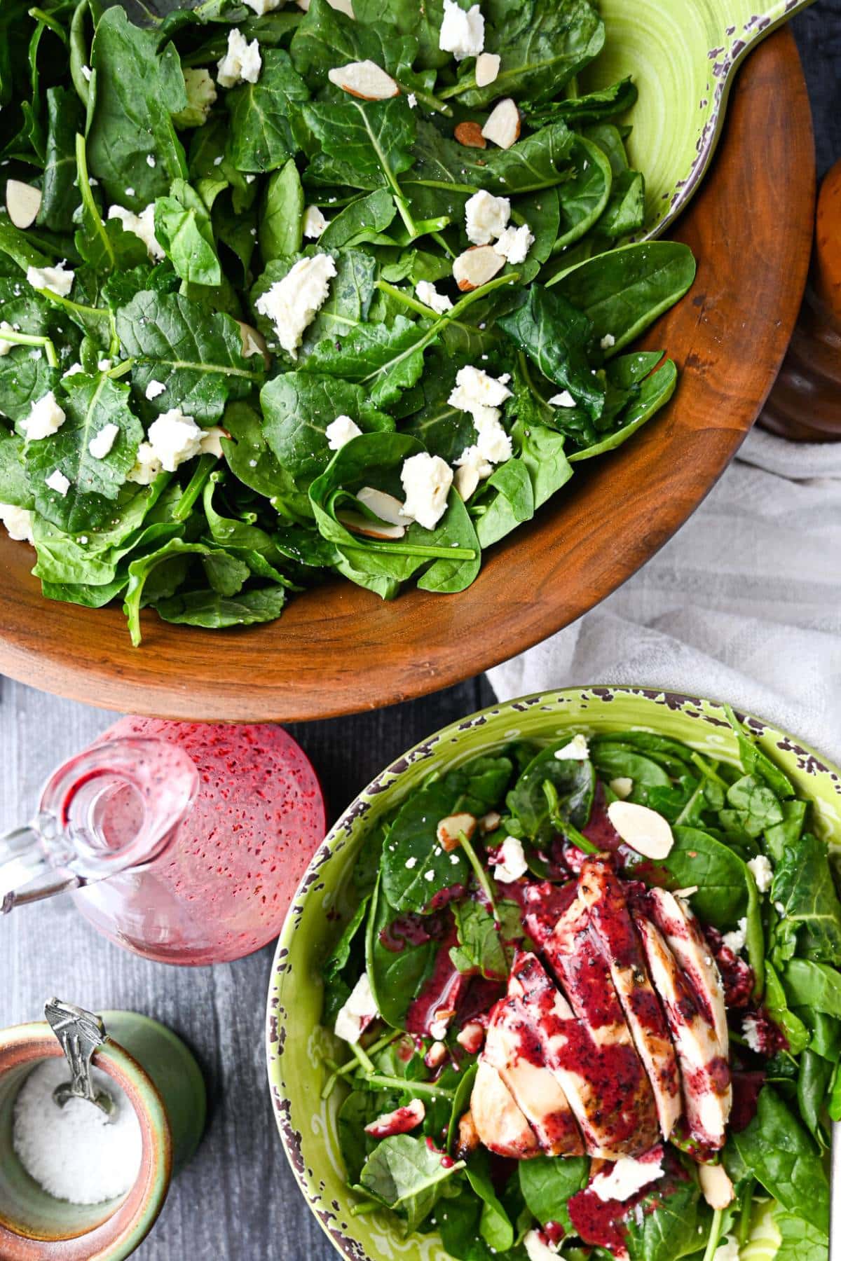 overhead photo of a wood serving bowl with prepared kale spinach salad, goat cheese, and almonds with a served bowl of salad topped with grilled chicken and blueberry vinaigrette dressing