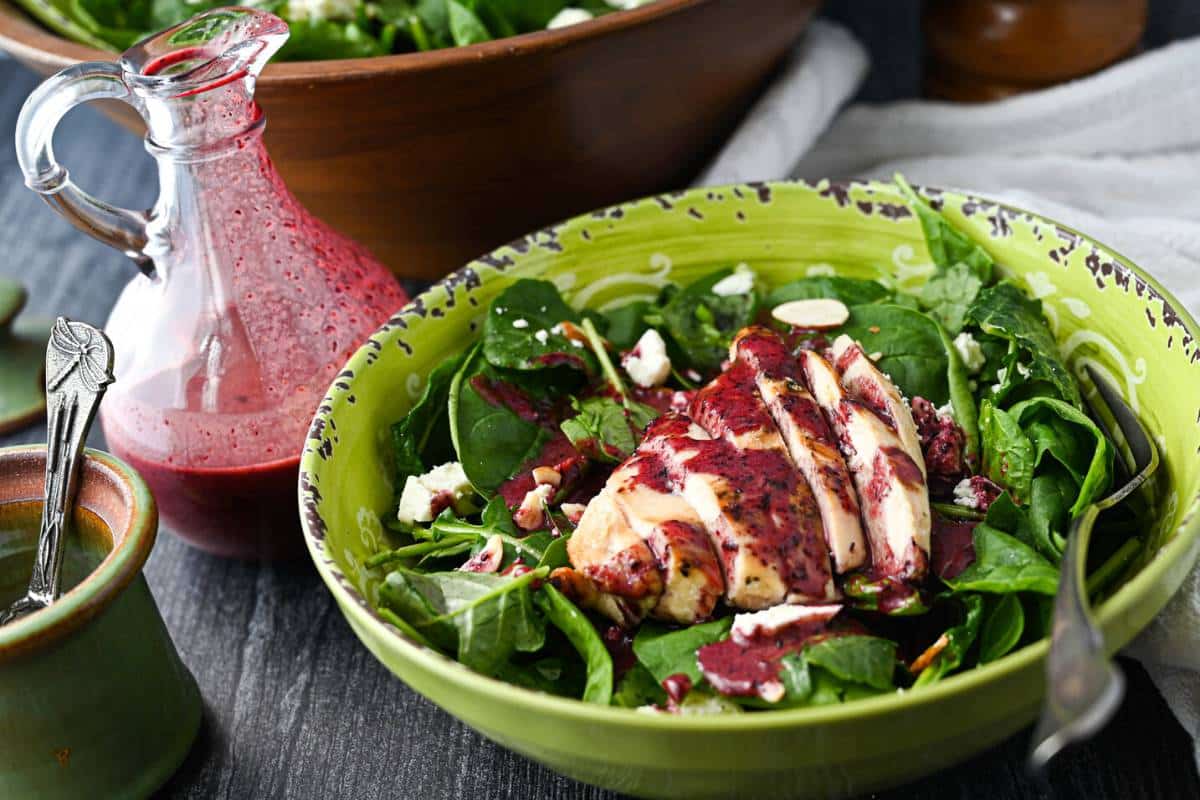 side view of a container of blueberry vinaigrette dressing with a bowl of kale spinach salad topped with a sliced grilled chicken breast