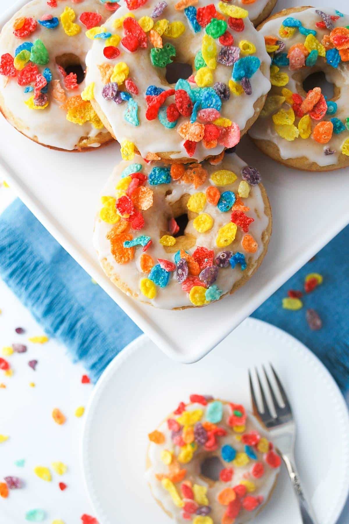 overhead photo of glazed vanilla protein donuts with Fruity Pebbles on a platter and a single donut with a fork on a plate below