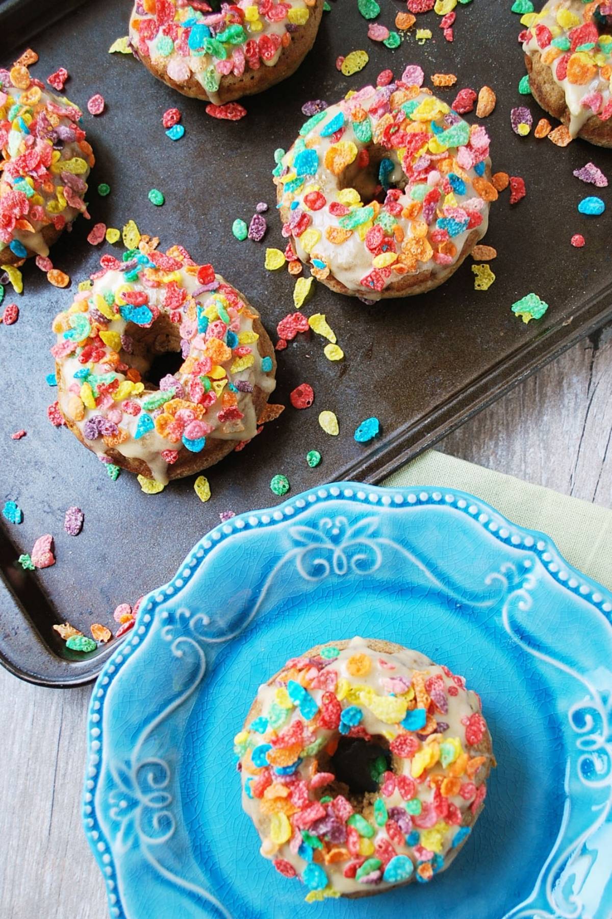 overhead photo of a vanilla protein donut covered in fruity pebbles on a blue plate with pan of donuts in the background