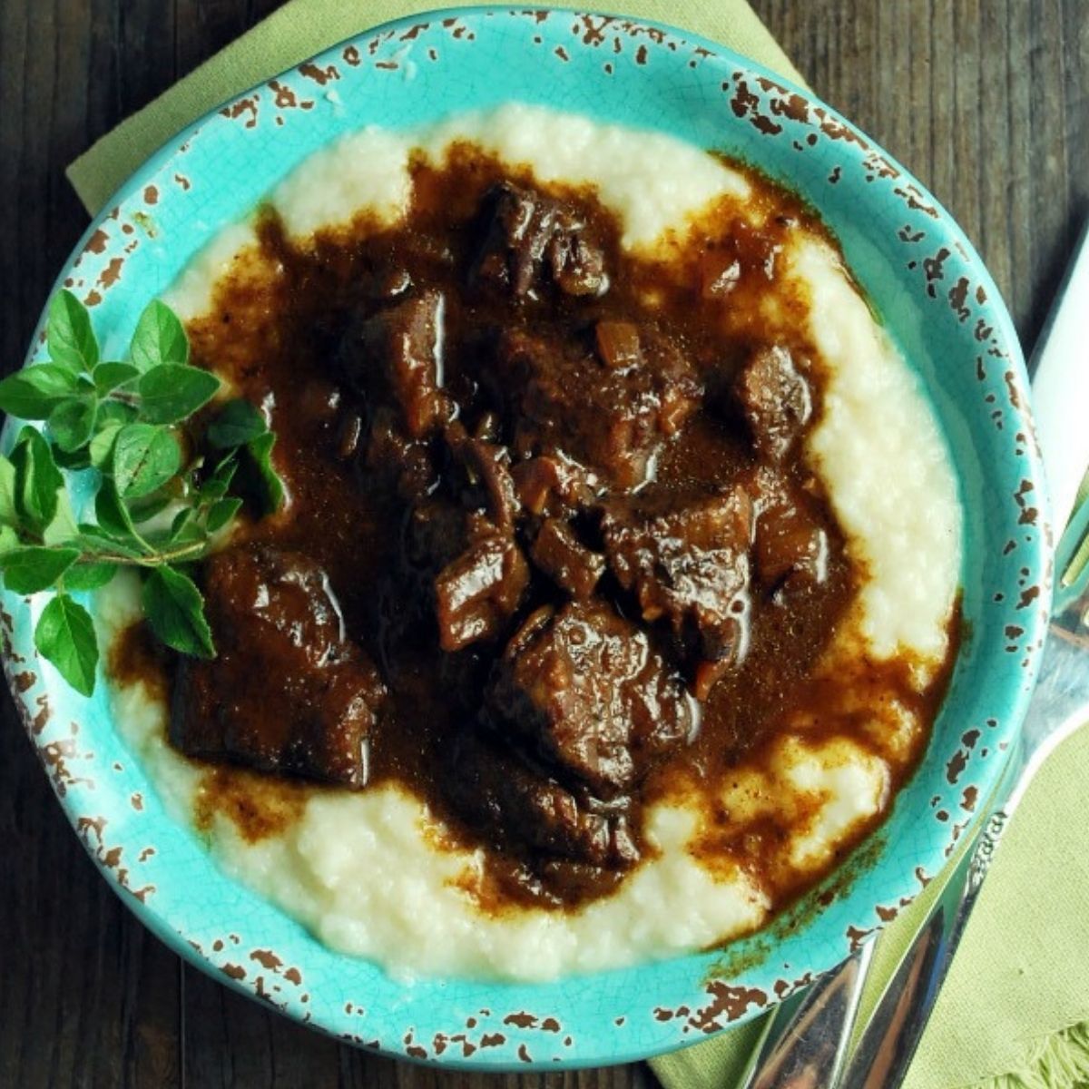 tender cooked beef with a rich gravy over cauliflower mashed potatoes in a blue bowl with a sprig of herbs