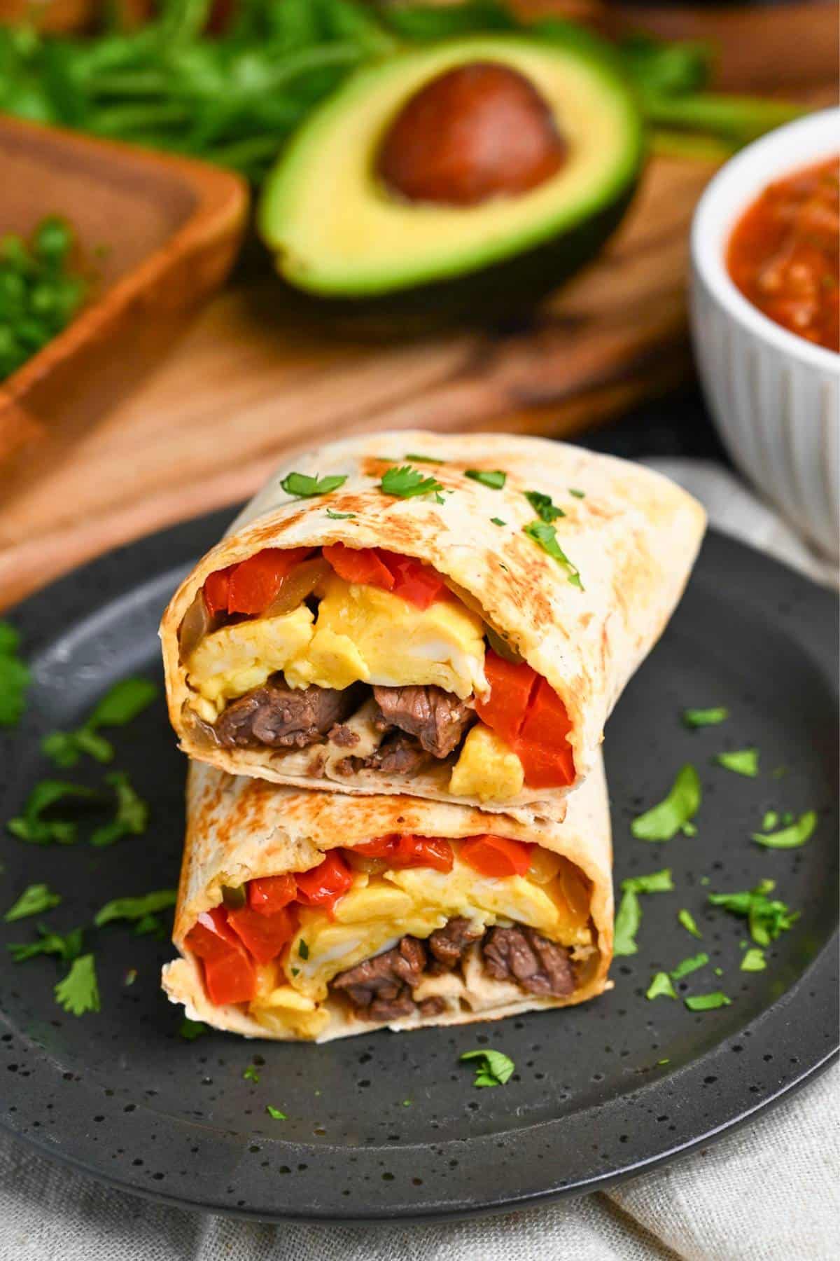 two burrito halves on a plate with a sliced avocado and salsa in the background