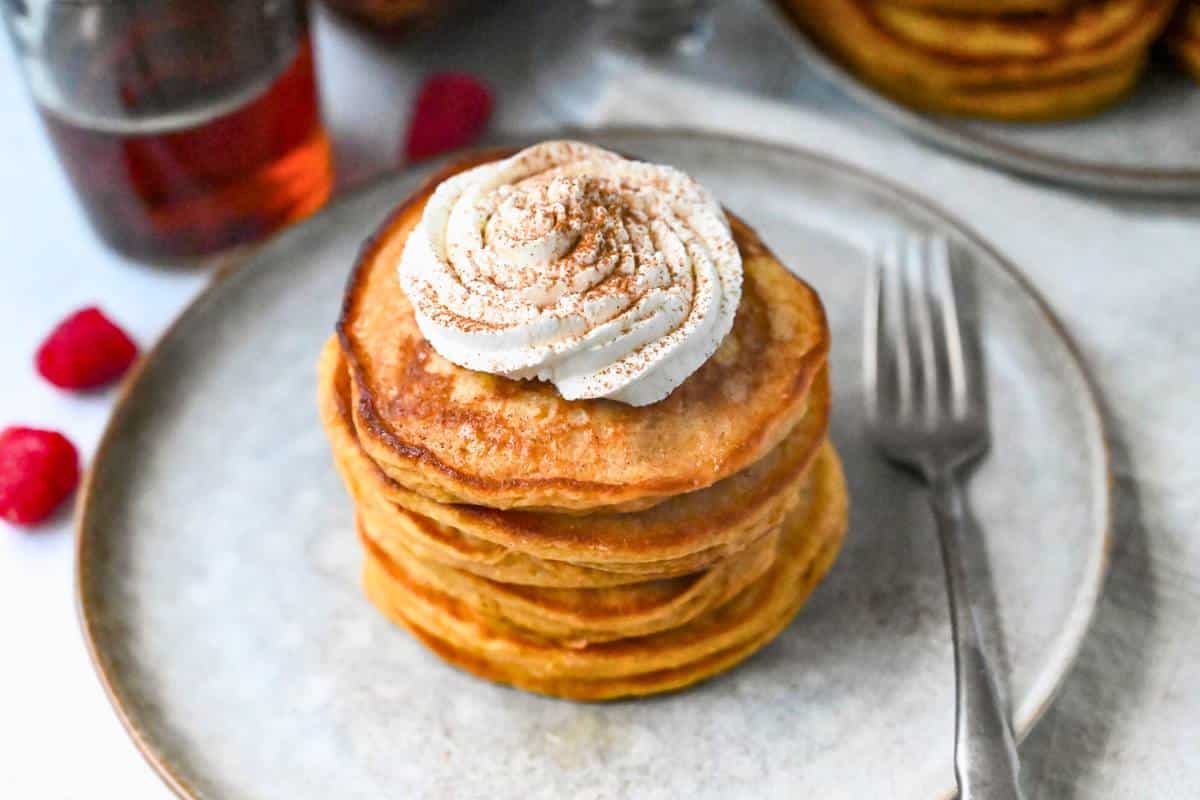 a plate of sweet potato kefir pancakes with whipped cream and cinnamon with a jar of syrup and fresh raspberries