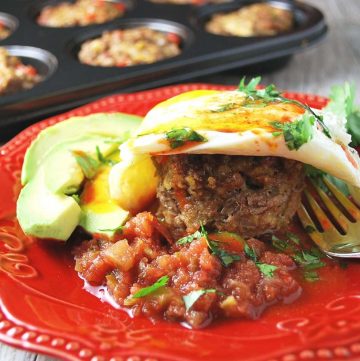 meatloaf muffin topped with a fried egg and parsley with sliced avocado and salsa on a red plate