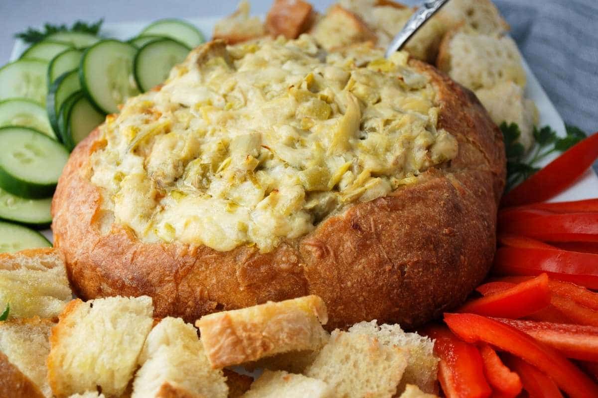 close up of artichoke dip in a bread bowl served with veggies and cubed bread