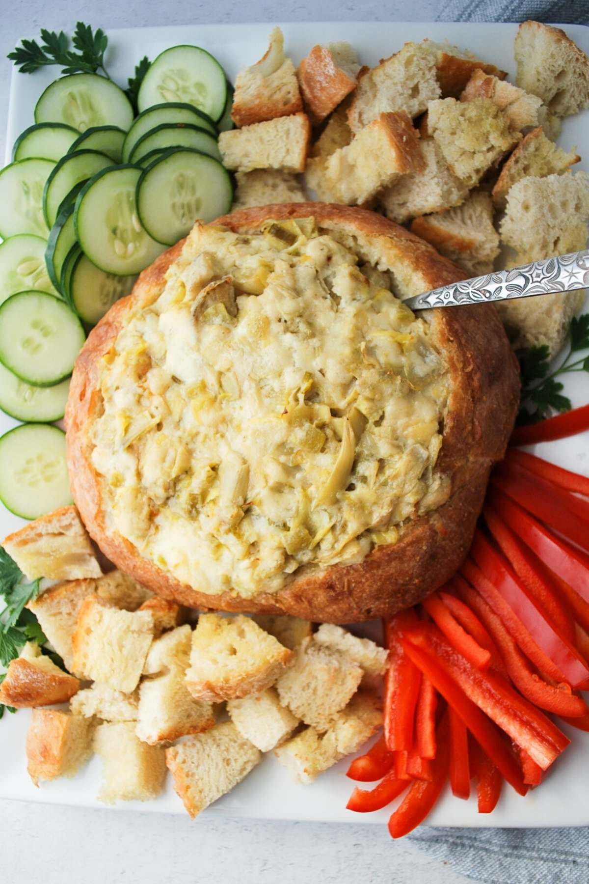 overhead shot of a baked bread bowl stuffed with artichoke dip and a platter of veggies and toasted bread cubes