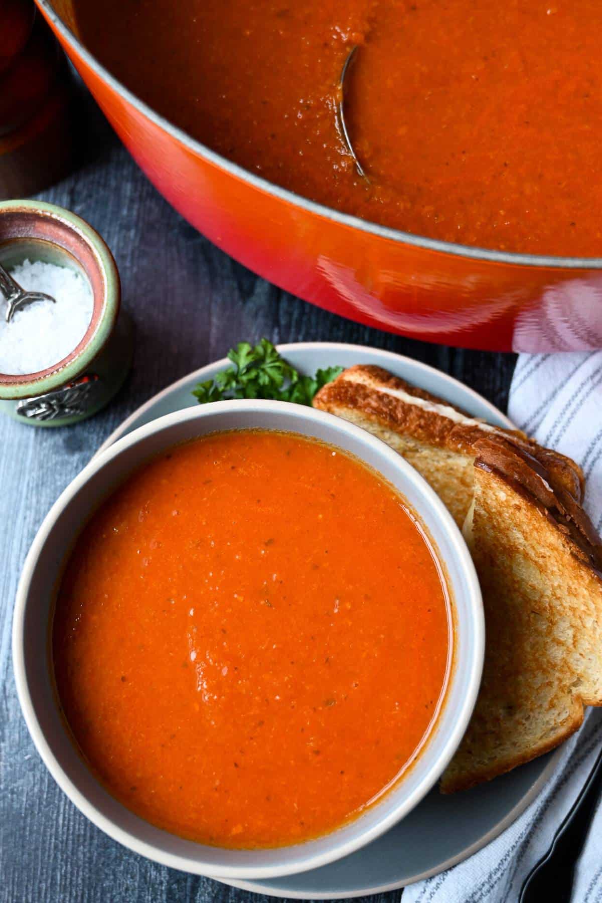 overhead photo of a dutch oven full of tomato bisque and a bowl of bisque with a grilled cheese sandwich