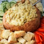 artichoke dip in a bread bowl surrounded by vegetables and bread cubes