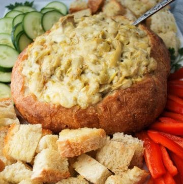 artichoke dip in a bread bowl surrounded by vegetables and bread cubes