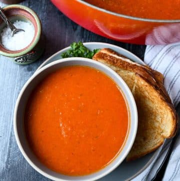 a bowl of tomato bisque with a grilled cheese on a plate with salt and a napkin next to it