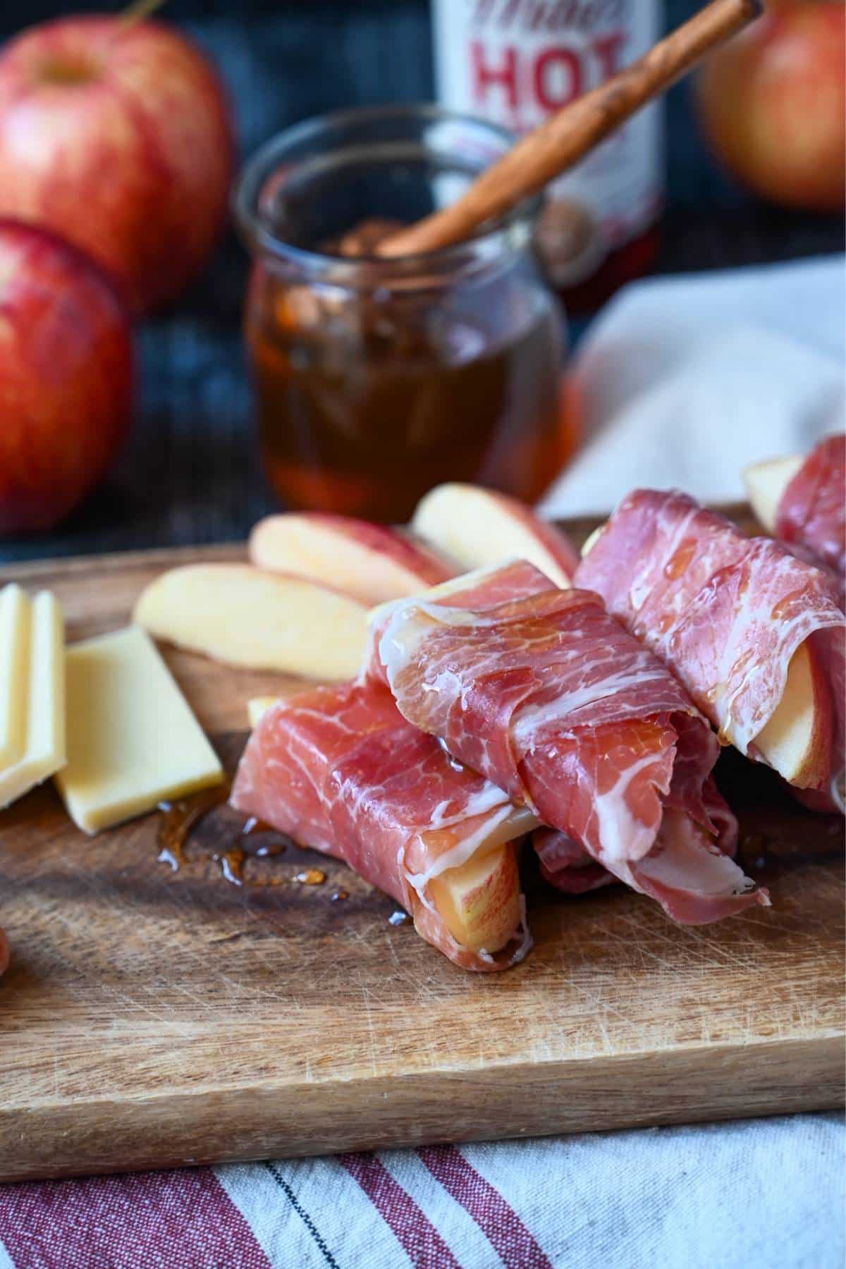 apple and cheese slices wrapped with prosciutto ham with honey drizzled on top. Jar of honey and apples in the background