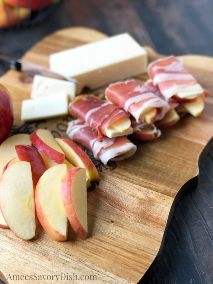 An apple sitting on top of a wooden cutting board, with Prosciutto wrapped apple slices and cheese