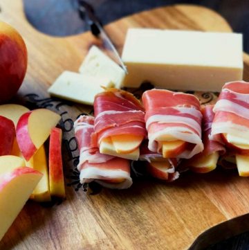 sliced apples and sharp cheddar cheese wrapped with slices of proscuitto ham on a cutting board with a block of cheese in the background