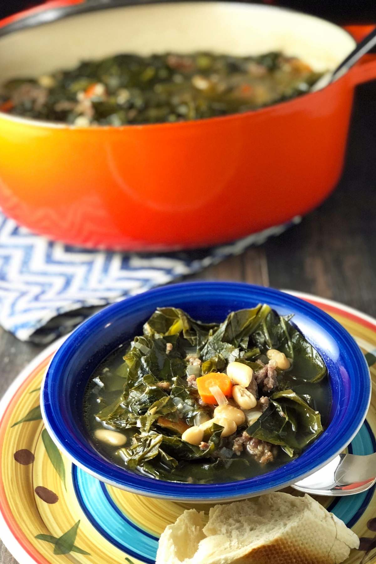 photo focused on a bowl of sausage soup with collards and white  beans with a blurred pot of soup in the background