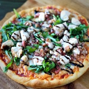 a whole pizza on a cutting board topped with arugula, diced chicken, pancetta, and balsamic glaze