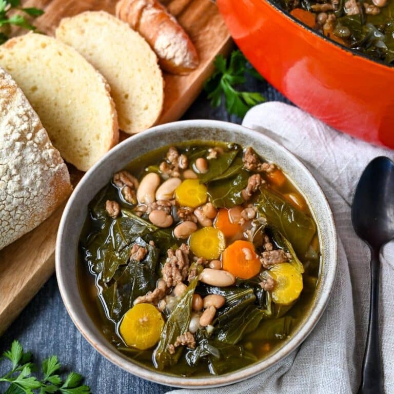 a bowl of collard green soup with Italian sausage and sliced fresh bread on a cutting board and an orange dutch oven of soup